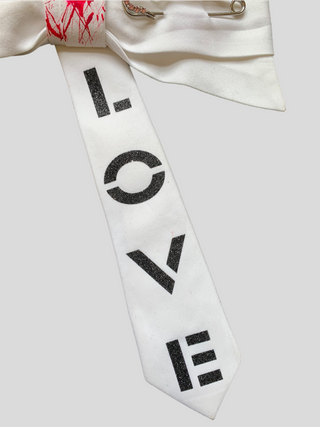 Closeup of White bowtie with the word Love painted in black, red splatter paint on knot and crystal kilt pin