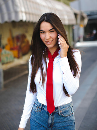 Woman wearing a white shirt, red tie, and blue jeans while on the phone outdoors.