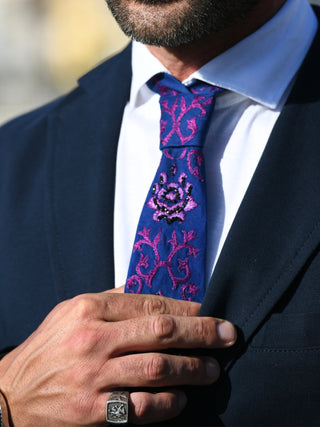 Male model holding his purple embroidered and beaded necktie against a dark suit and white shirt with a  visible pinky ring 