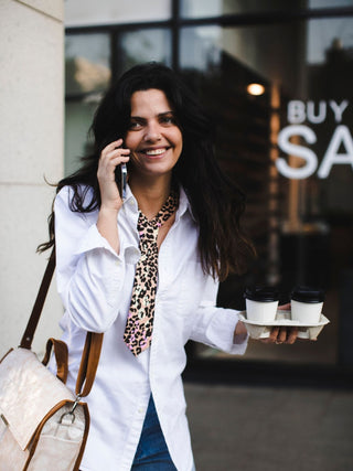 Model talking on a phone, holding coffee cups and a handbag, wearing a leopard print petite tie over a white shirt, with a store window in the background.