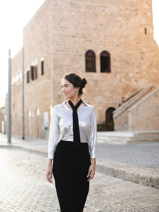 Model wearing a black classic necktie over a blouse and black pants in front of a brick building.